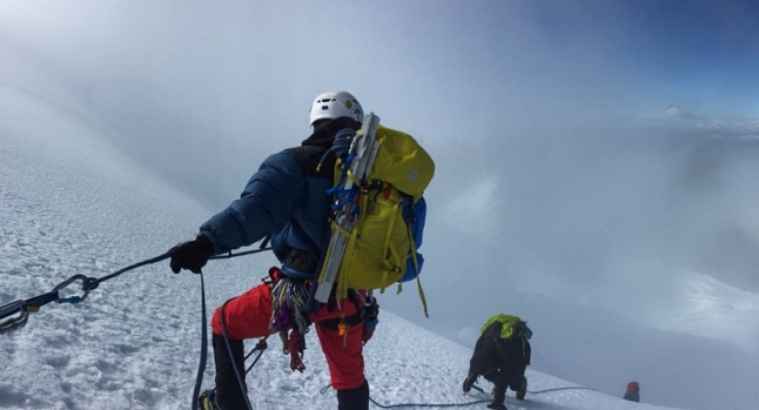 A group of mountaineers wearing safety gear are connected by a rope. The person in the foreground looks down the snowy slope to two others below them.